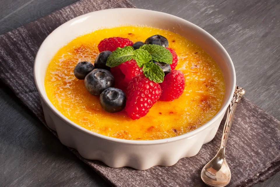 Crème brûlée topped with fresh raspberries, blueberries, and mint leaves, served in a white ramekin with a spoon on the side