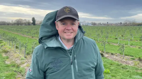 BBC A man wearing a green baseball cap and green rain jacket is standing in a field with hundreds of small plants in the background.