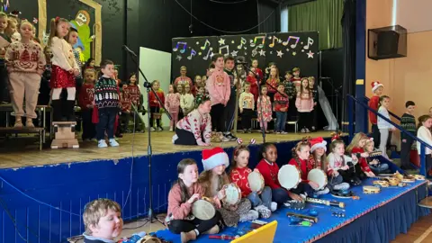 Group of children all together on stage with Christmas hats on. They are standing with instruments. 