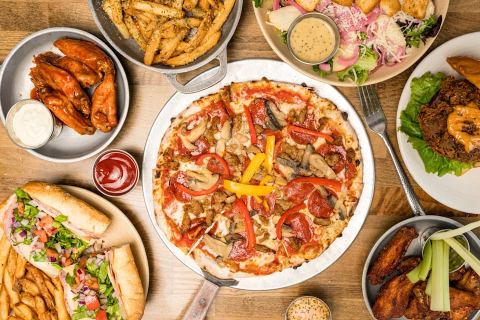 Table filled with a variety of foods: pizza, wings, fries, a sandwich, a salad, and a breaded dish, accompanied by dipping sauces