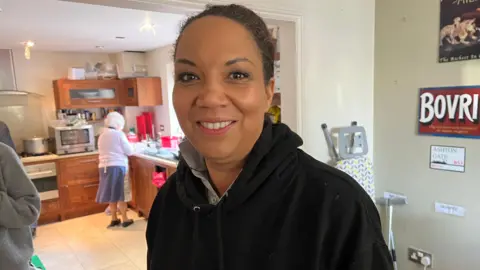 BBC Caz Goddard is smiling, wearing a black hooded jumper while stood near a kitchen. An elderly woman is pictured doing the washing up behind her.