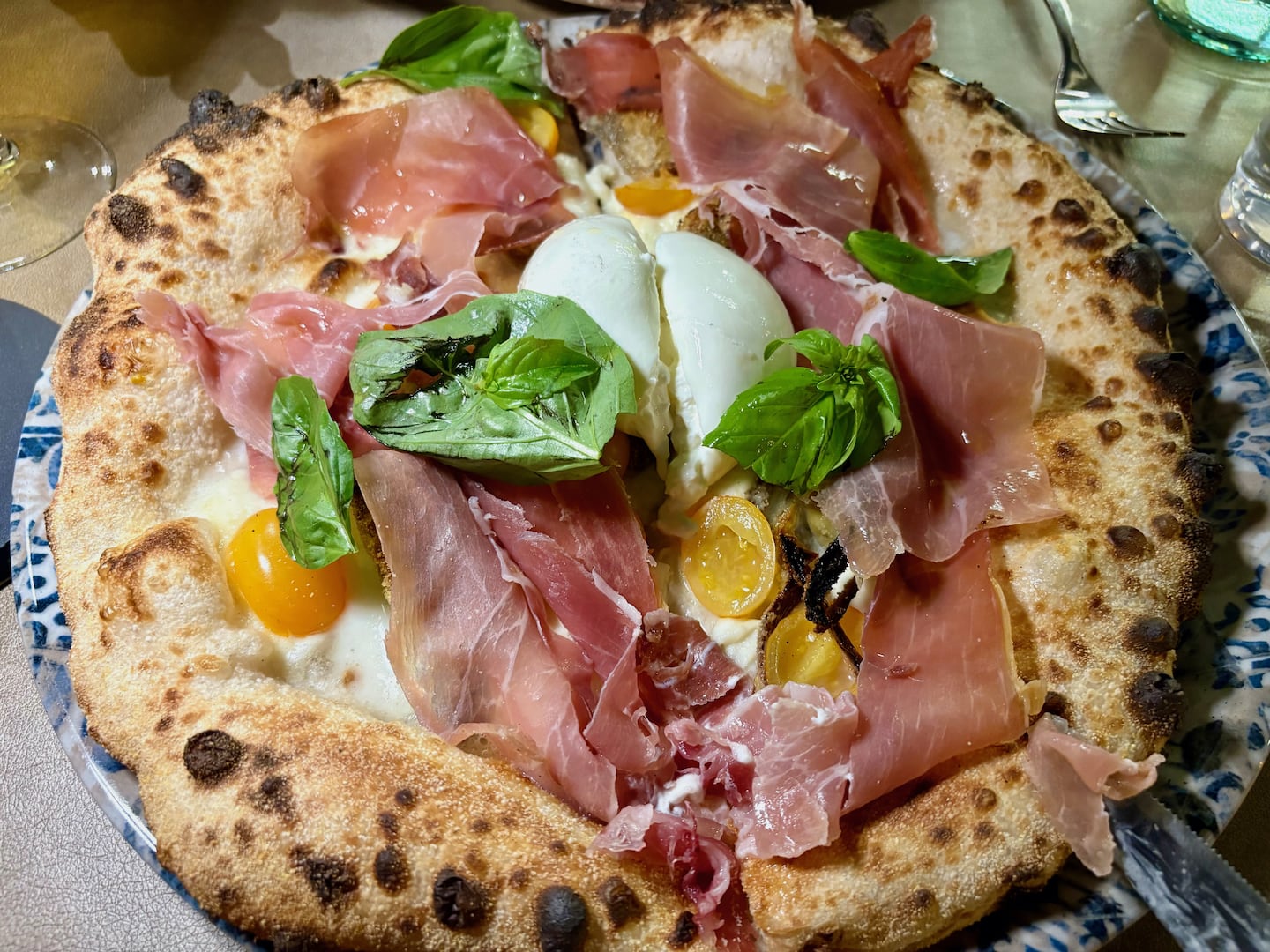 At Bollicine in Palermo. a fire-singed, pillowy crust pizza is topped with salty prosciutto, sweet yellow cherry tomatoes, creamy Buffalo mozzarella, and earthy, nutty artichokes.