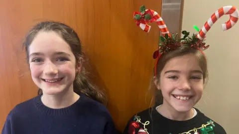 Two girls smiling at the camera. One of the girls is wearing a candy cane headband. Behind them is an oak door. 