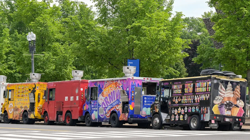Food trucks lined up in a row, each covered with vibrant graphics and menu items, parked on a street with trees in the background