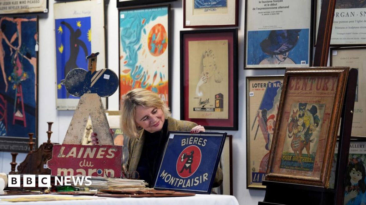 A woman sits at a table holding and looking at a enamel sign. All around her are prints and signs from the 20th century.