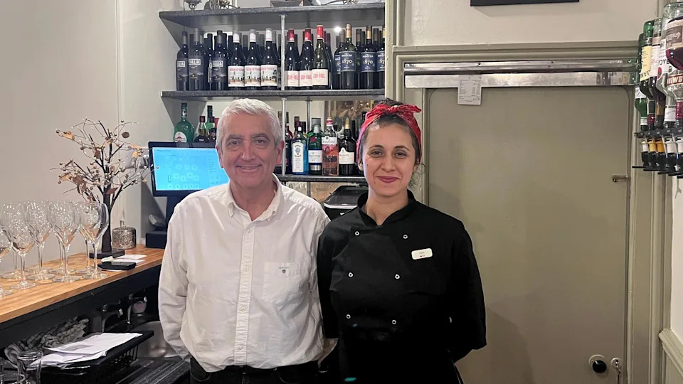 A man and a woman stand behind a bar and smile at the camera.