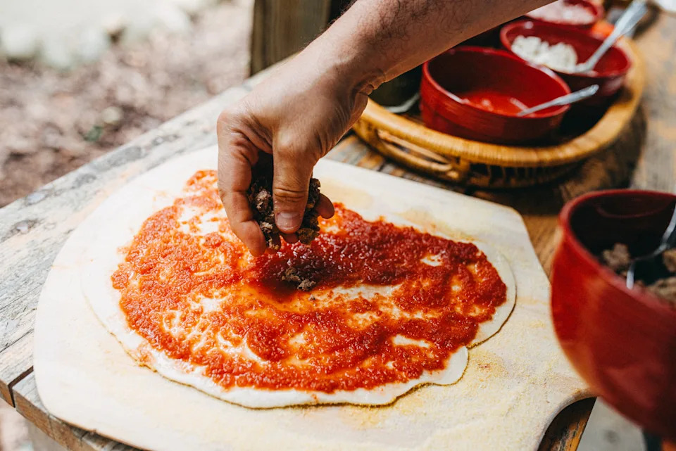 Person applying sauce on pizza dough with bowls of ingredients nearby on a wooden table