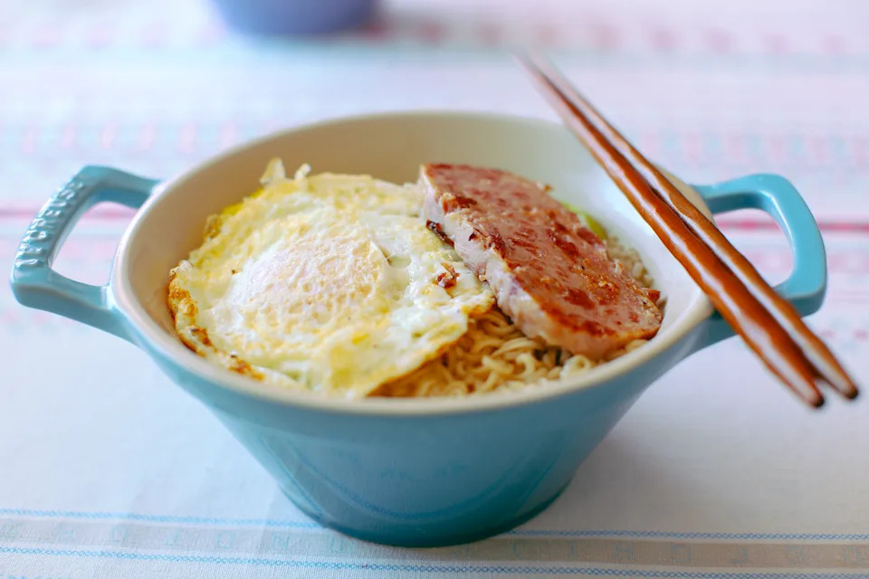 Bowl of instant noodles topped with a fried egg and a slice of grilled meat, with chopsticks resting on the rim