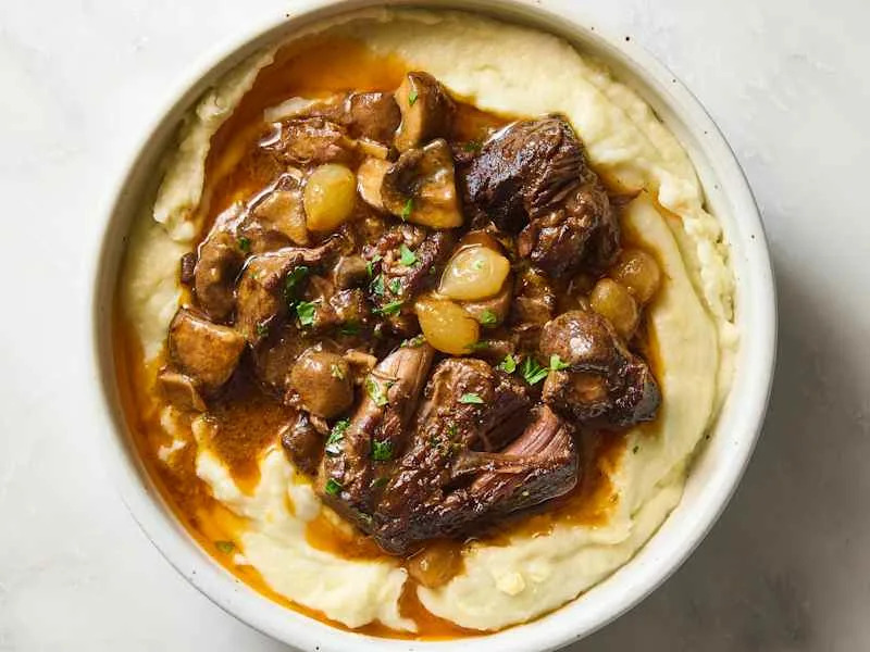Beef stew with mushrooms and pearl onions served over creamy mashed potatoes, garnished with parsley.