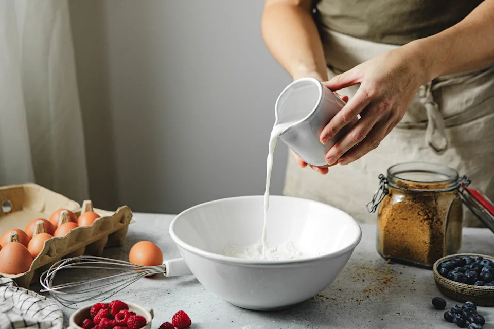 Midsection of woman pouring milk on wheat flour in bowl. Female is standing in kitchen is preparing pancake with ingredients around.