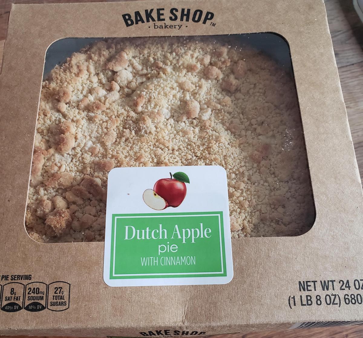 A boxed Dutch apple pie with cinnamon is shown through a clear window in the packaging. The label features an image of an apple and green text. The box displays nutritional information and the net weight of 24 oz.