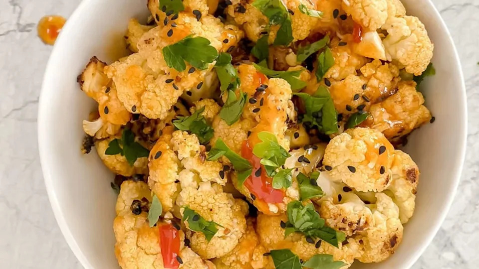 Overhead shot of prepared bang bang cauliflower in white bowl garnished with fresh parsley and black sesame seeds atop marble countertop.