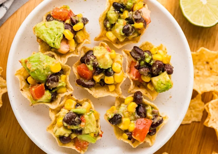 Tortilla chips topped with black beans, corn, avocado, and diced tomatoes are arranged on a white plate.