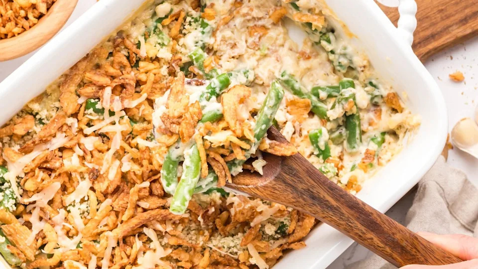 Partially scooped green bean casserole in a white ceramic baking dish with a wooden spoon, showing crispy onion topping and creamy interior.