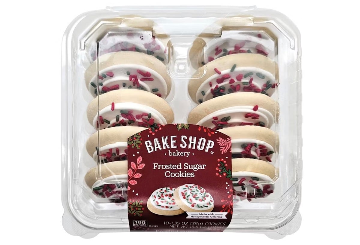 A clear plastic container holds twelve frosted sugar cookies with white icing and red and green sprinkles. The label reads “Bake Shop bakery Frosted Sugar Cookies” and features festive holiday designs.