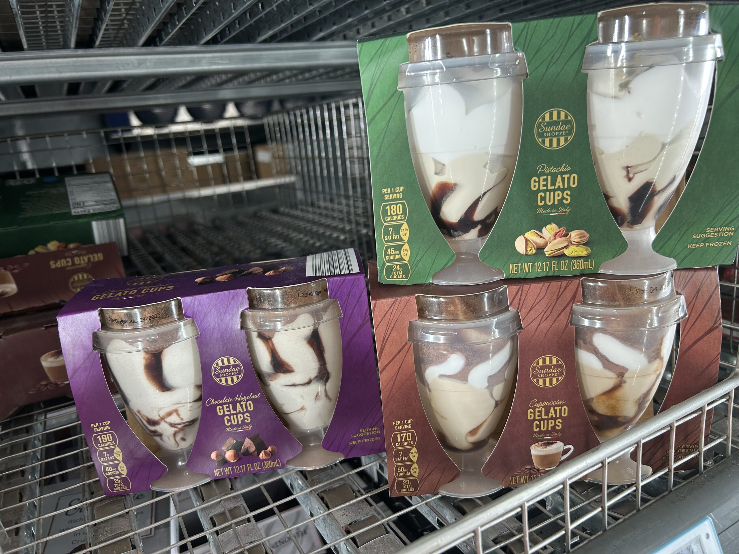 Boxes of Sundae Shoppe gelato cups in clear plastic containers are displayed on a metal freezer shelf. Flavors include pistachio (green box) and triple chocolate (purple box).