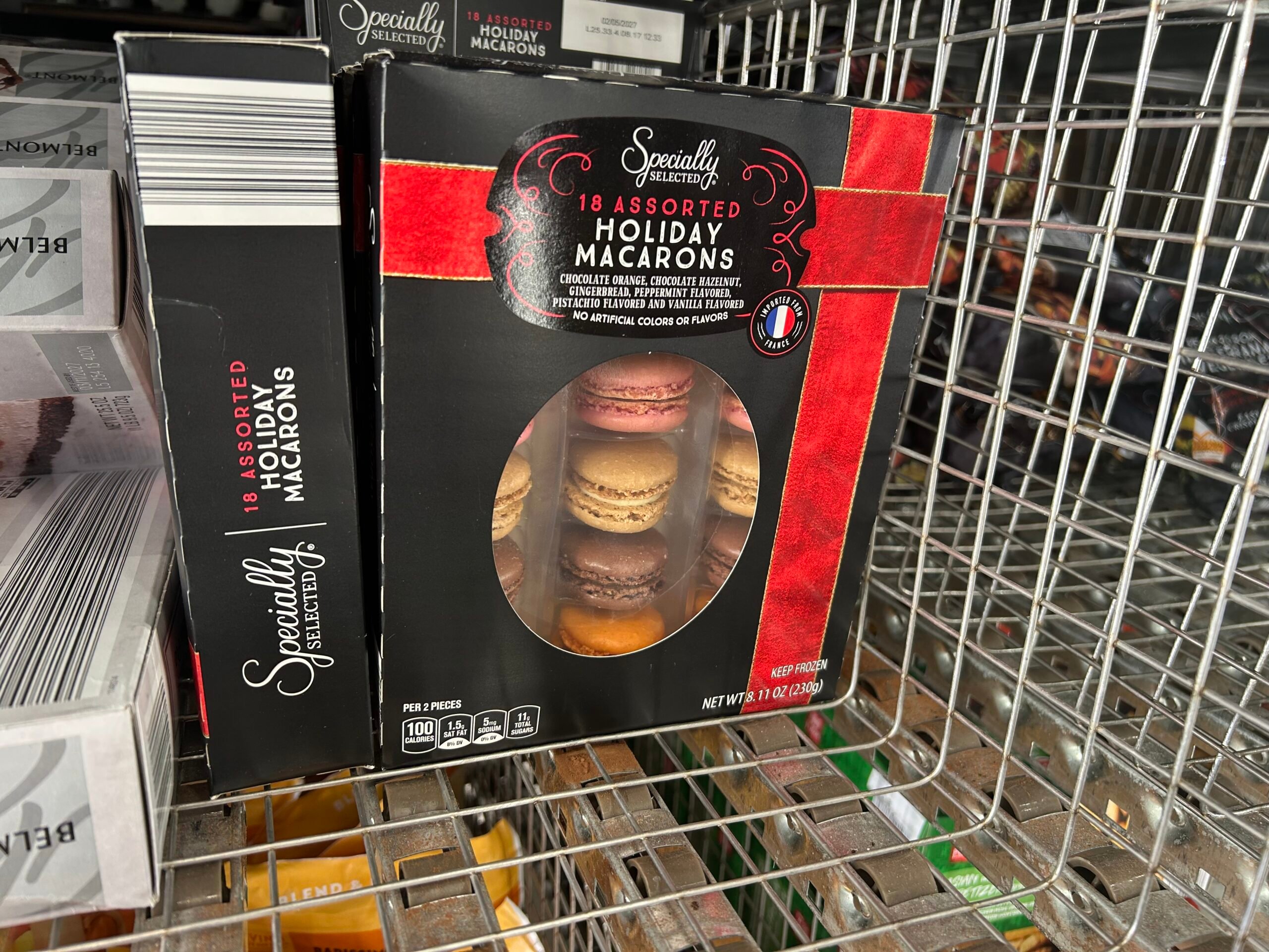 A black and red box labeled "18 Assorted Holiday Macarons" sits on a wire shelf. The box has a clear window showing various colored macarons inside. Other boxes and store items are visible nearby.