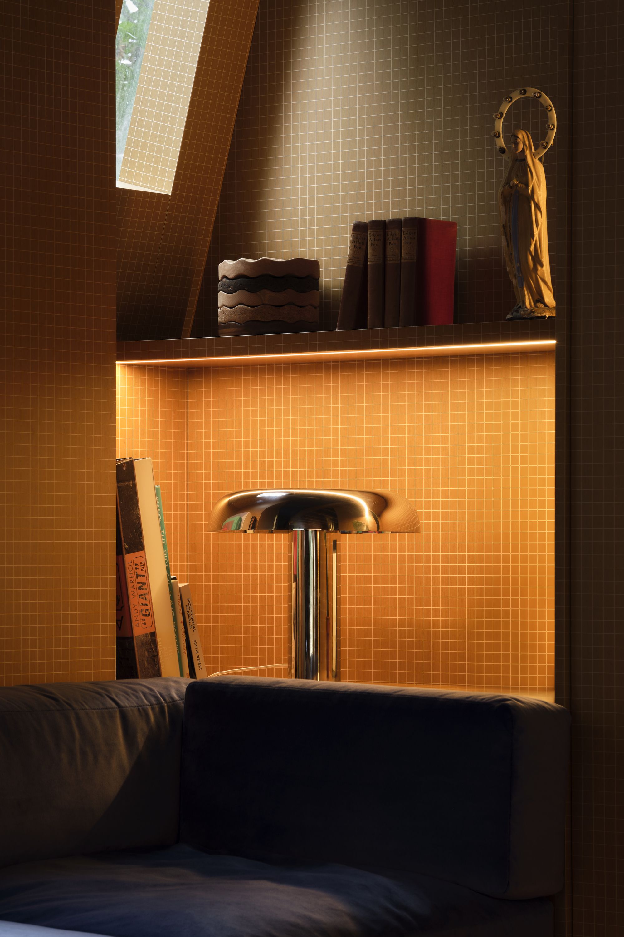 the corner of a navy blue sofa against a wall made of yellow subway tiles. There is a built-in shelf with books and a chrome mushroom lamp on it.