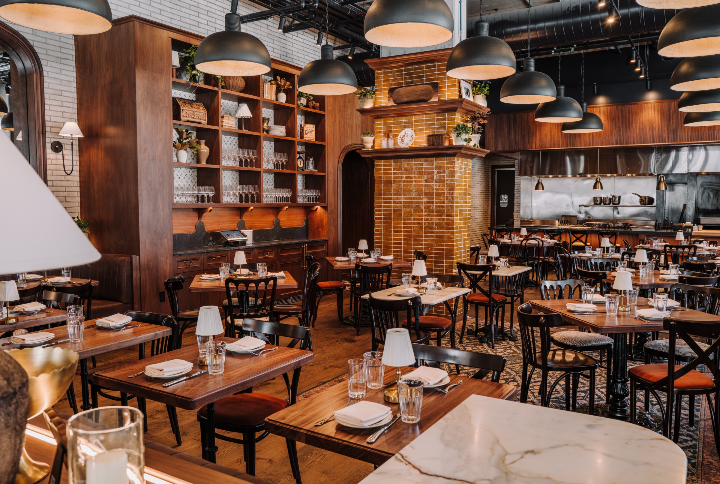 A dining area with two- and four-top tables, hanging lights, and shelves lined with glasses and decorations.