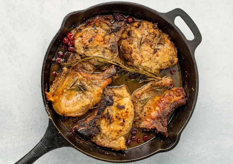 Four cooked pork chops with rosemary and cranberries in a cast iron skillet, viewed from above on a light background.