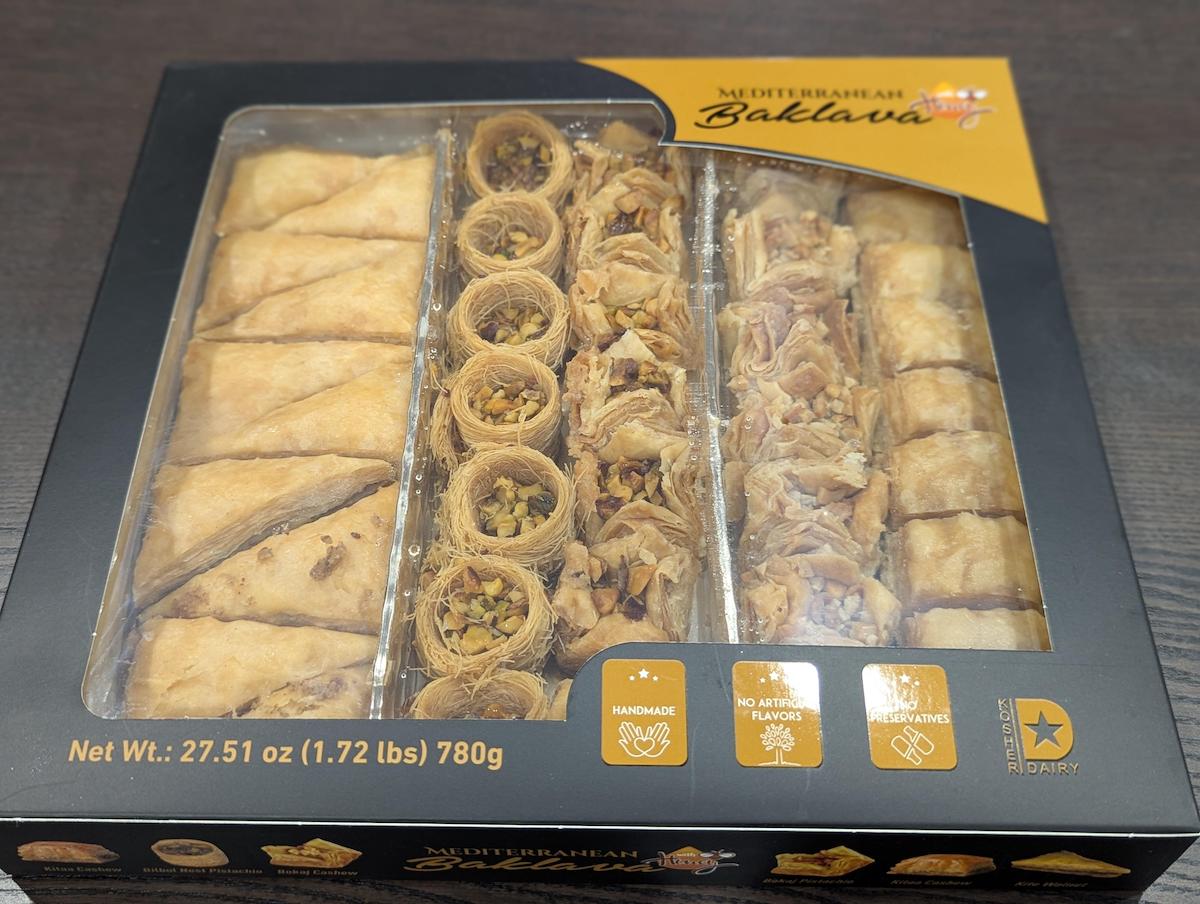 A box of assorted Mediterranean baklava with three types neatly arranged in rows, visible through a clear window on the packaging. The box highlights that it's handmade, with no artificial flavors or preservatives.