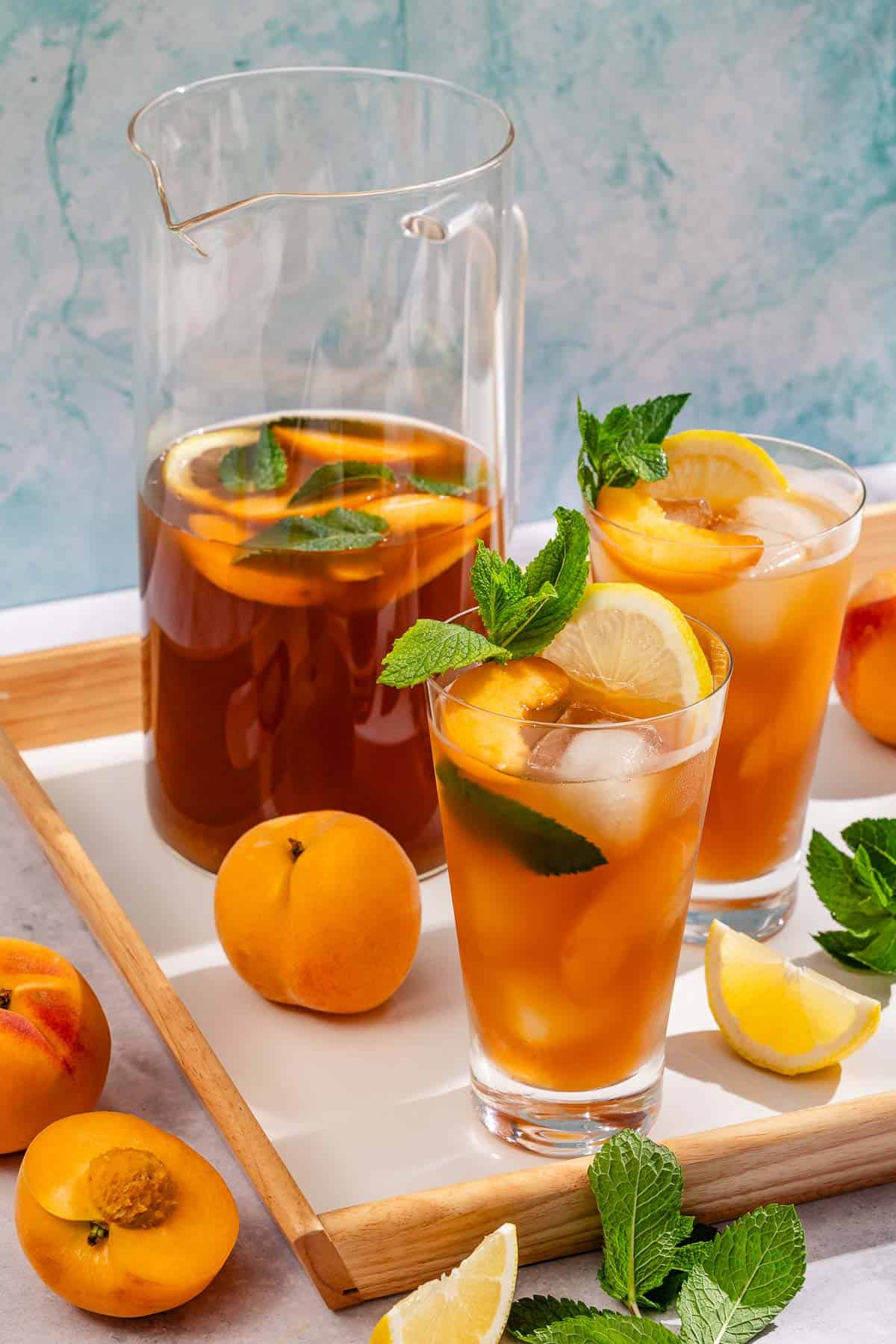 Two glasses of peach tea garnished with mint and peach and lemon wedges. Next to this is a pitcher of the tea, peaches, lemon wedges and fresh mint.