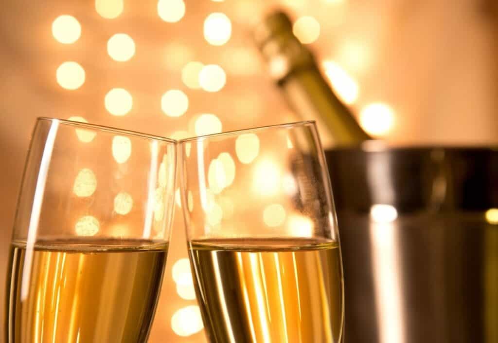 Two glasses of champagne clinking together with a bottle of champagne in an ice bucket and warm lights in the background.