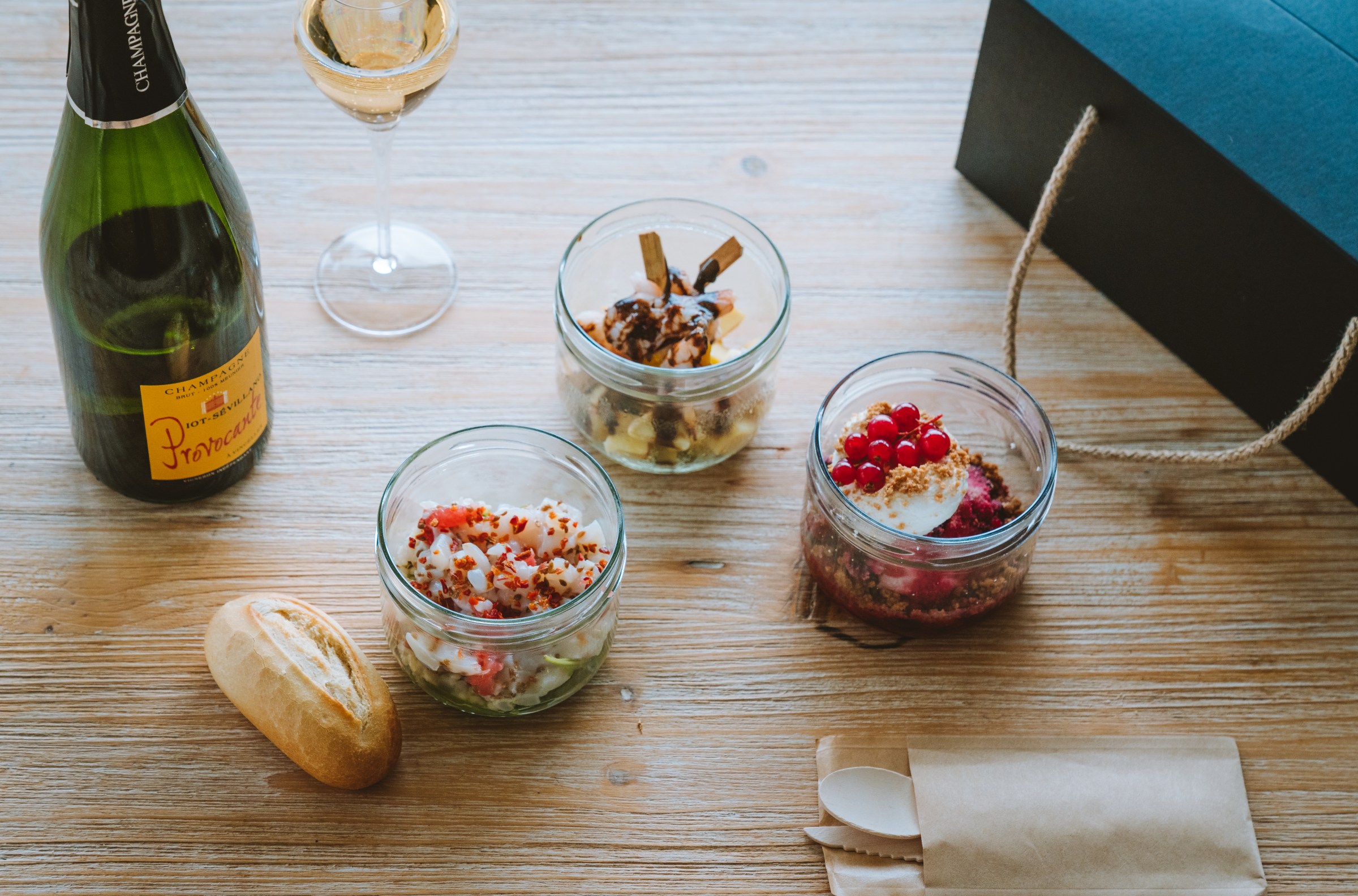 Three small glass jars hold gourmet dishes next to a bottle of champagne on a wood table.