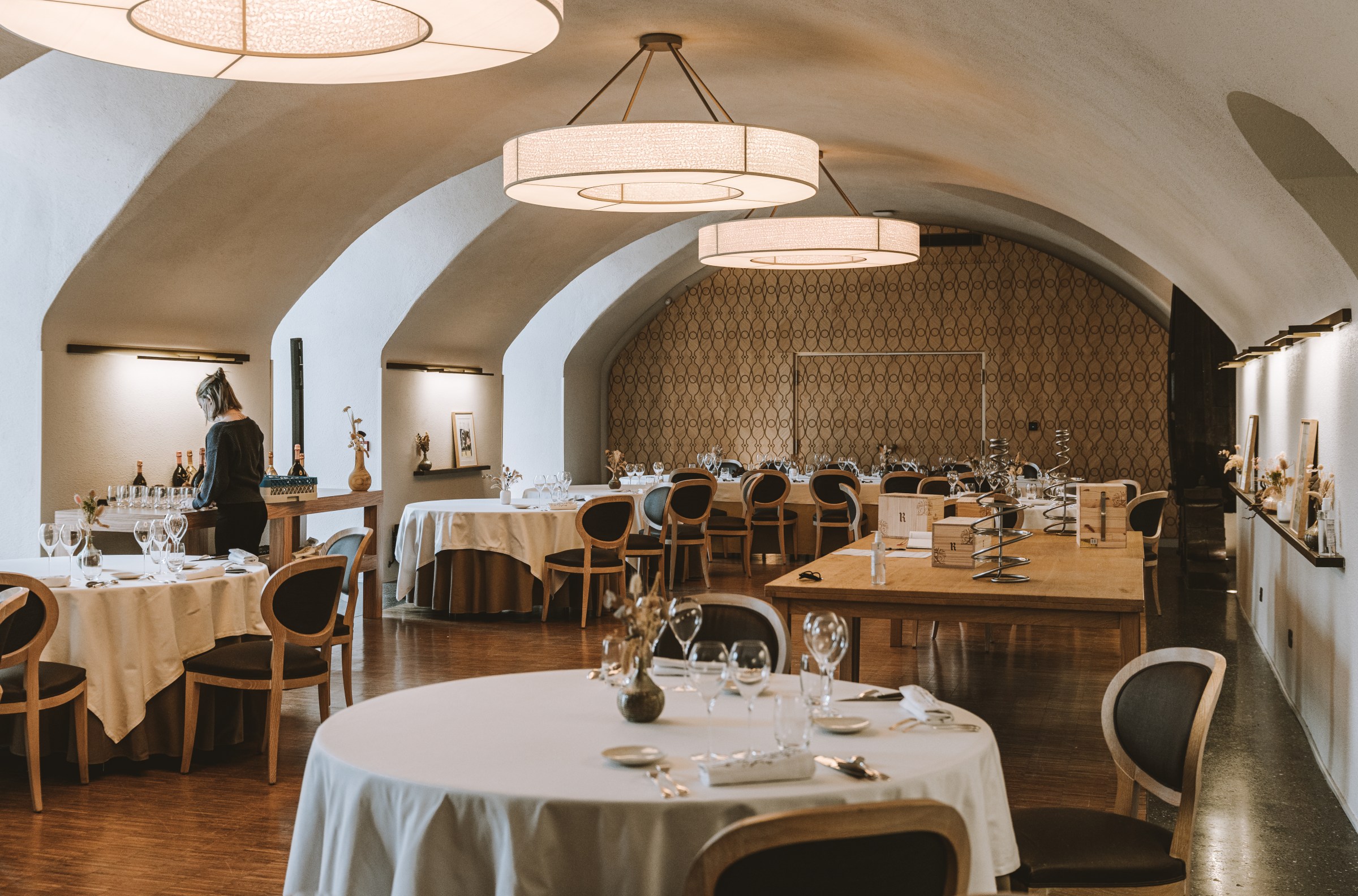 A waitress pours wine in a dining rom with white cloth-covered tables and high arched ceilings. 