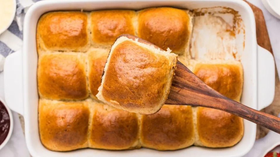 Golden-brown rye dinner rolls baked in a white rectangular dish, with one roll lifted out using a wooden spatula to reveal its soft interior.
