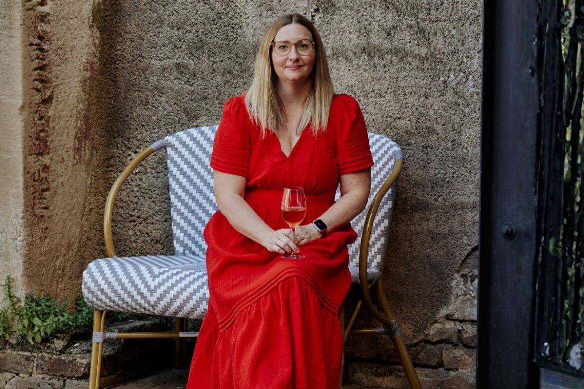 Kristen Hall, in a red dress, sits on a patterned bench outdoors, holding a glass of wine against a textured stone wall.