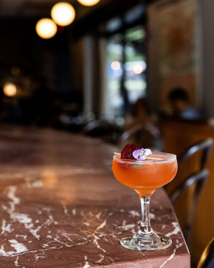 A coupe glass with an orange cocktail garnished with a slice of strawberry and a small flower sits on a marble bar counter in Birmingham’s La Fête.