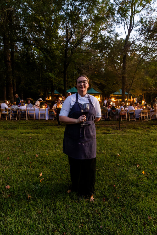 A chef standing in front of a dining event, holding a glass of wine.