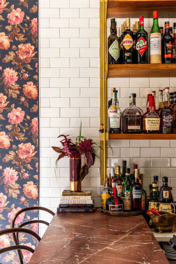 Marble bar counter with assorted liquor bottles on wooden shelves, a floral wallpaper on the left, and a vase with red flowers on a stack of valuable items.