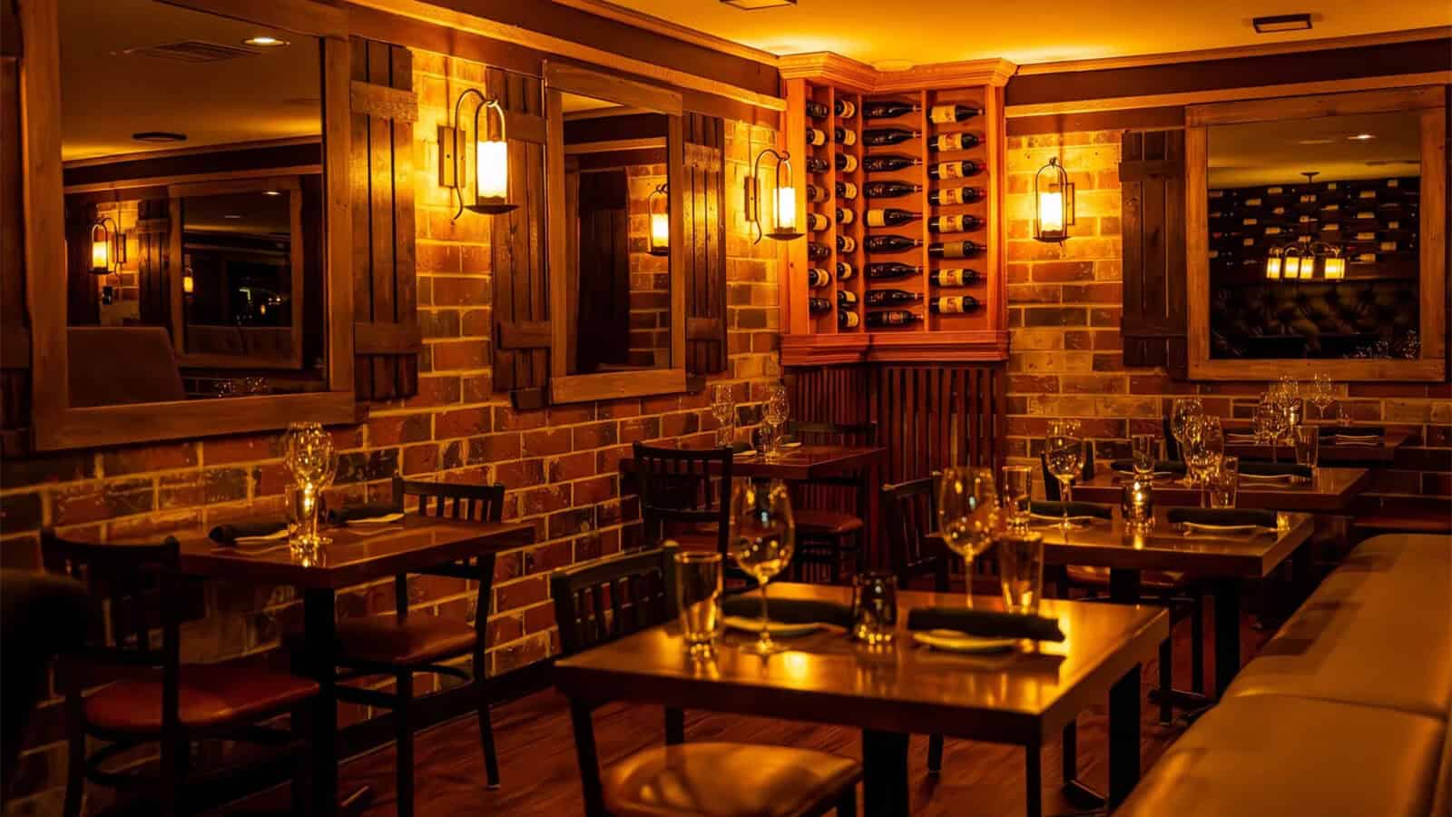 A warmly lit restaurant dining area at Sansonina Ristorante Italiano features wooden tables, brick walls, mirrors, wine bottles on display, and neatly arranged glassware and settings—a true celebration of counter culture dining.