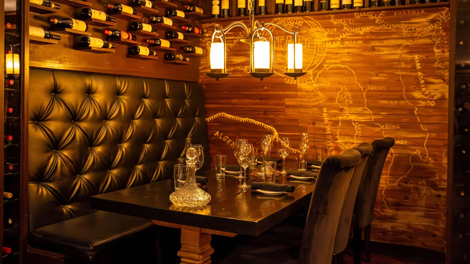 A dimly lit booth at Sansonina Ristorante Italiano features a black tufted bench, wine bottles on racks, set tableware, and a wooden wall adorned with a map design—capturing the essence of an inviting Italian restaurant.