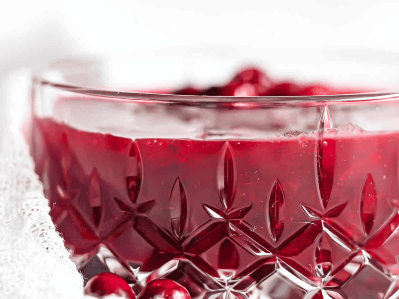 A close-up of a crystal bowl filled with red cranberry sauce, with whole cranberries visible inside and around the bowl.