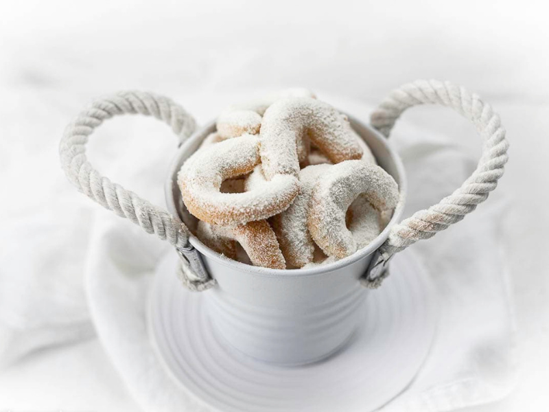 A white metal bucket with rope handles holds crescent-shaped cookies coated in powdered sugar, placed on a white plate with a white cloth background.