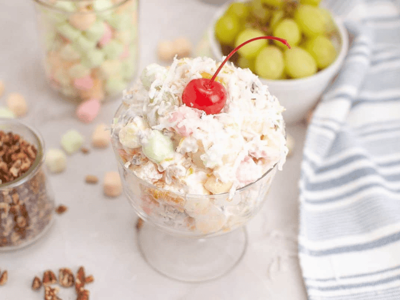 A glass dish filled with ambrosia salad, topped with a maraschino cherry, sits on a table with grapes, marshmallows, nuts, and a striped cloth nearby.