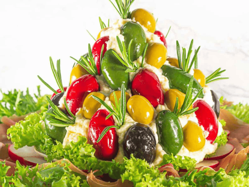 A mound of creamy cheese spread is decorated with multicolored olives and sprigs of rosemary, surrounded by leafy greens and slices of cured meat.