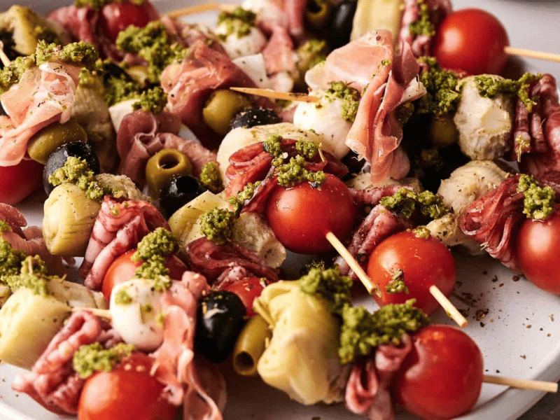 A plate of antipasto skewers with cherry tomatoes, mozzarella, olives, artichoke hearts, cured meats, and a drizzle of green pesto.