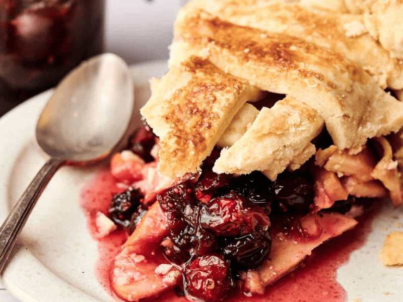 A slice of mixed berry pie with a golden crust sits on a plate next to a spoon, with fruit filling spilling out.