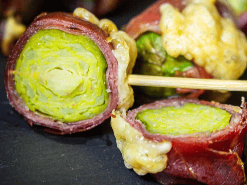 Close-up of Brussels sprouts wrapped in prosciutto and topped with melted cheese, skewered on a wooden stick.