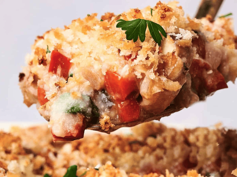 A close-up of a spoon lifting a portion of a baked breakfast casserole dish containing vegetables, topped with a golden breadcrumb crust and garnished with a parsley leaf.