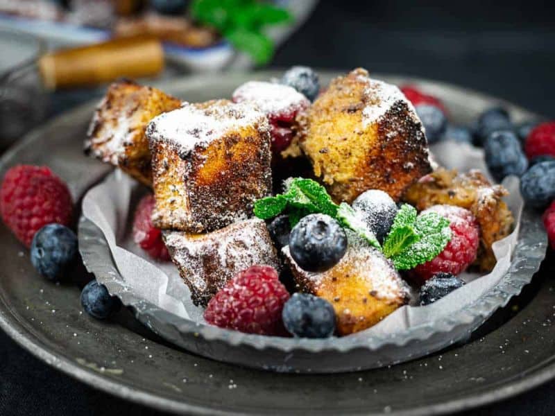 Cubes of French toast dusted with powdered sugar, served with fresh raspberries, blueberries, and a sprig of mint on a dark plate.