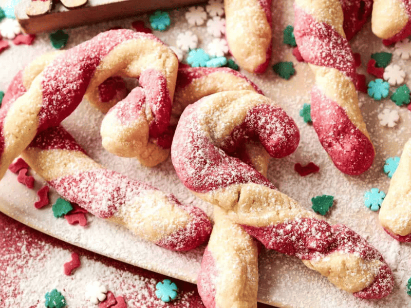 Twisted red and white candy cane cookies dusted with powdered sugar, surrounded by decorative snowflake and tree-shaped sprinkles.
