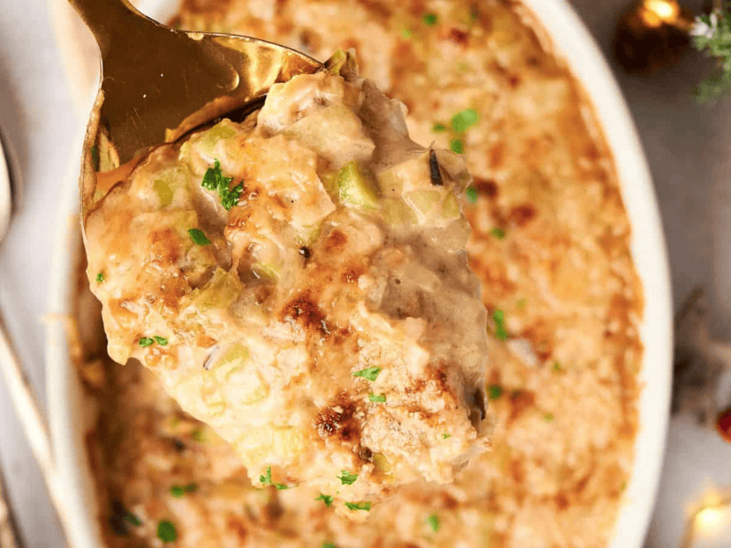 A close-up of a spoon scooping a portion of creamy baked casserole topped with herbs, with a dish of the casserole and a spoon in the background.