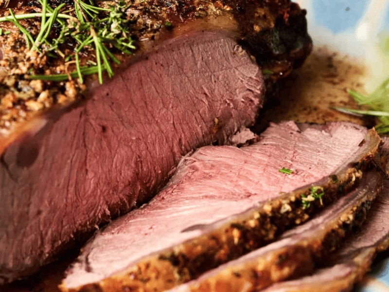 Close-up of a cooked roast beef with a herb crust, partially sliced to show the juicy, medium-rare interior.