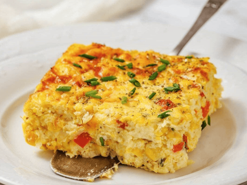 A square slice of corn pudding casserole with eggs, red bell pepper, and chives sits on a white plate with a serving spoon underneath.