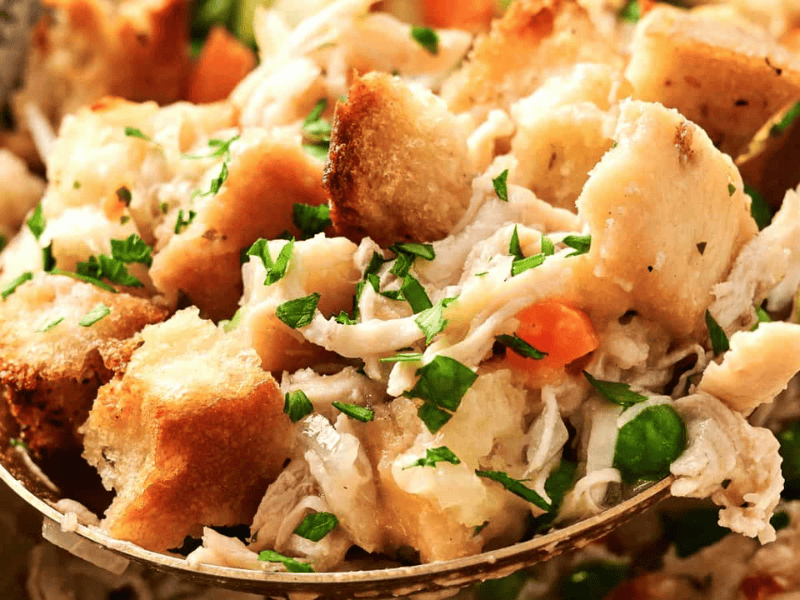 A close-up of a spoonful of chicken stuffing, showcasing croutons, shredded chicken, peas, carrots, and sprinkled herbs.